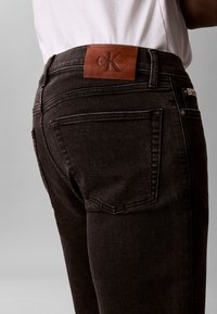 Μαύρο τζιν από denim με καφέ δερμάτινο λογότυπο Calvin Klein στη μέση, φορεμένο με λευκό πουκάμισο, εμφανίζεται από τη μέση πίσω έως το μέσο του μηρού.