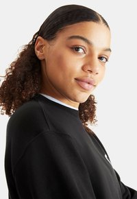 Black sweatshirt with a crew neck, textured fabric, and a white inner collar. Features round logo accent on the chest. Model's face slightly visible.