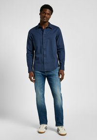 Mörkblå långärmad skjorta med krage, slim fit och logodetalj, i kombination med ljusa denimjeans och avslappnade vita sneakers.