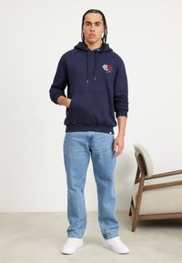 Sudadera navy con bolsillo canguro y bordado floral; combinada con jeans azul claro y zapatillas blancas; ajuste casual y relajado.