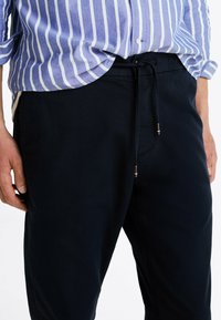 Pantaloni blu navy in tessuto testurizzato, con vita regolabile tramite cordoncino e dettagli in metallo. Abbinati a una camicia a righe blu chiaro.