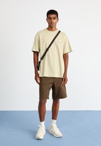 Beige cotton t-shirt with a simple graphic design, paired with brown shorts. White socks and cream sneakers complete the outfit.