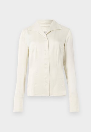 Blouse à manches longues de couleur blanc cassé, avec un large col et une taille cintrée, présentée sur un fond clair et uni.