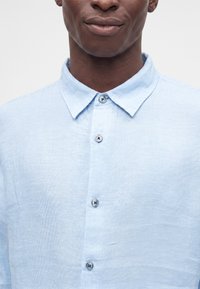 Homme portant une chemise bleu clair boutonnée avec un col, montré du cou jusqu'au milieu de la poitrine, sur un fond clair uni.