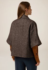 Bruine herringbone jas met een kraag, brede manchetten en korte mouwen, gelaagd over een bruine blouse. Zachte textuur en gestructureerd ontwerp.