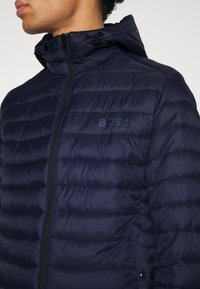 Marinblå vadderad jacka med dragkedja och huva med snörning. Har horisontell kviltning och en diskret logotyp på bröstet.