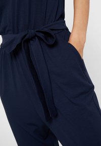 Gros plan sur une personne portant un pantalon bleu marine avec une ceinture en tissu nouée en nœud papillon et une main placée dans une poche latérale.