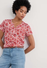 Camicia a maniche corte rossa e bianca con un motivo floreale, scollo a V e una vestibilità comoda, abbinata a jeans a vita alta di un blu chiaro.