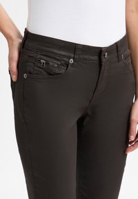 Donkerbruine jeans met een slim fit, voorzien van een ritszak, metalen accenten en subtiele stikseldetails. Gemaakt van een soepele, rekbare stof.