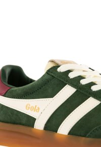 Zapatillas de ante verde con rayas blancas y acentos en marrón. Presentan una lengüeta crema, un logo dorado y una suela translúcida marrón.