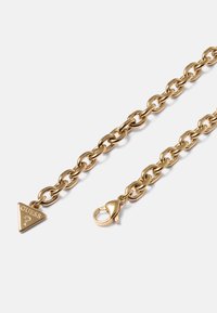 Goldkette mit ovalen Gliedern, ausgestattet mit einem dreieckigen "Guess"-Logo-Anhänger und einem Karabinerverschluss vor schlichtem weißen Hintergrund.