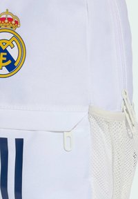 Bijela torba napravljena od tkanine s plavim i žutim logotipom Real Madrida, mrežasta bočna džep i zatvarač s bijelim detaljima.