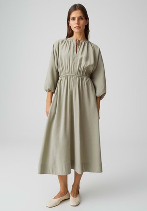 Vrouw staand, gekleed in een losse midi-jurk van lichtolijfgroen met lange pofmouwen en beige instapschoenen, tegen een effen achtergrond.