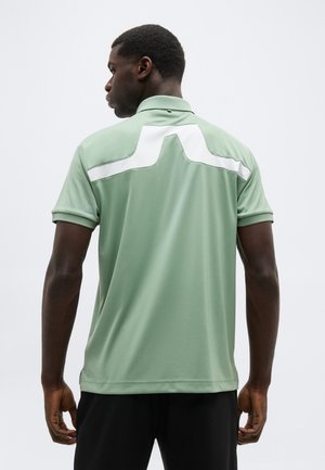 Man met een lichtgroen poloshirt met een witte geometrische streep over de bovenrug, staand met de rug naar de camera, tegen een effen achtergrond.