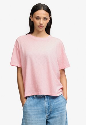 Mujer con cabello largo y oscuro que lleva una camisa de manga corta suelta de color rosa claro y vaqueros de color azul claro con las manos en los bolsillos sobre un fondo blanco.