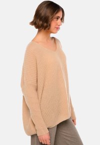 Pull en tricot beige avec une coupe ample, décolleté en V et texture côtelée. Manches larges et ourlet arrière légèrement plus long. Design décontracté.