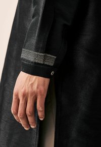 Black garment with a textured surface, showcasing a cuff with silver horizontal stripes and a metal snap button detail.