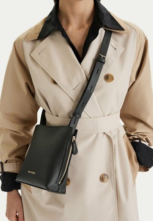 Beige trench coat with black collar, worn with black crossbody bag featuring gold zipper and adjustable strap.