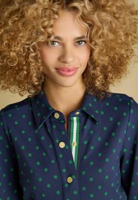 Jūras zila krekls ar zaļiem punktiņiem, ar pogām priekšpusē, zelta pogas un svītrainu zaļo un balto akcentu uz pogu sloksnes.