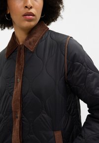 Črna prešita jakna z ribanim rjavim kordurojnim ovratnikom in gumbnim zapiranjem. Ima ukrivljene šive in gladko teksturo.