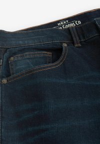 Dunkelblaue Denim-Jeans in Nahaufnahme, die die Vordertasche, Münztasche mit oranger Naht und Knopfdetails sowie ein teilweise sichtbares Markenlabel im inneren Bund zeigen.