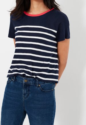 Kvinde iført navyblå t-shirt med hvide horisontale striber og rød halsudskæring, kombineret med mørkeblå denimjeans, hænderne bag ryggen.