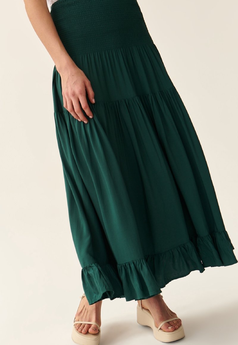 Femme portant une jupe à volants vert foncé à plusieurs niveaux avec un ourlet à volants et des sandales compensées beiges sur un fond clair uni.