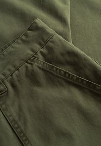 Tkanina olivno zelene barve z gladko teksturo, z žepom in šivanimi detajli vzdolž šivov. Opazna je subtilna sijajnost.