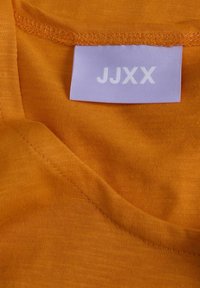 Tela naranja con una textura suave, que presenta un escote en V. Una etiqueta de color púrpura claro con "JJXX" es visible en el lado interior.