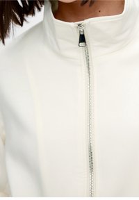 Giacca bianca con zip e colletto alto, con zip argentata e texture liscia. La vista ravvicinata mette in evidenza i dettagli delle cuciture.