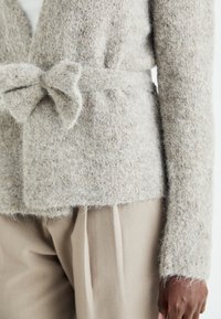 Cardigan bej texturat cu un cordon, având un material moale și pufoș, cu mâneci lungi. Purtați peste pantaloni de culoare deschisă, cu o croială lejeră.