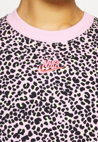 Rosa T-shirt med kort ärm som har ett svart och grönt leopardmönster, med en röd broderad Nike-logotyp på bröstet. Den är tillverkad av strukturerad tyg.