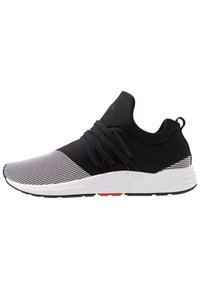 Schwarz-weißer Sportschuh mit einem gestrickten Obermaterial, strukturiertem Muster, elastischen Schnüren und einer dicken weißen Sohle mit roten Akzenten.