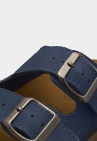 Μπλε σουεντ λουρί σανδαλιού με δύο ασημένιες μεταλλικές ρυθμιζόμενες αγκράφες και ορατές ραφές σε κοντινό πλάνο.