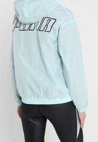 Ljust blå huva jacka med elastisk nederkant, med en stor svart "PUMA" logga på baksidan, tillverkad av lätt, något texturerat material.