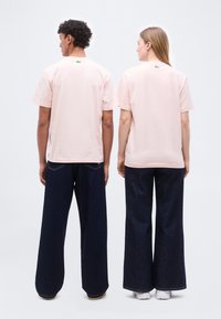 Δύο άτομα γυρισμένα προς τα πίσω, φορούν ανοιχτό ροζ μπλουζάκια και σκούρα τζιν με φαρδιά μπατζάκια, με φόντο ένα απλό ανοιχτό τοίχο.