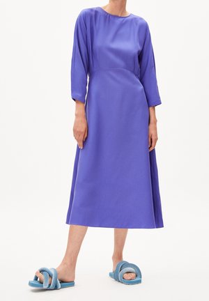 Femme portant une longue robe violette à manches trois-quarts et des sandales slides bleues avec de larges brides rembourrées.