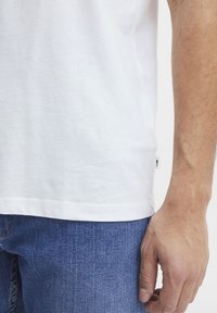 Camiseta de algodón blanca con escote redondo, mangas cortas y una pequeña etiqueta en el dobladillo. Combinada con jeans de mezclilla azul.