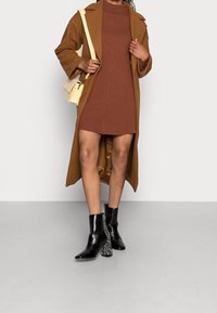 Robe marron à côtes, superposée avec un long manteau marron. Bottines noires avec un design texturé et des talons à motifs. Sac bandoulière jaune.