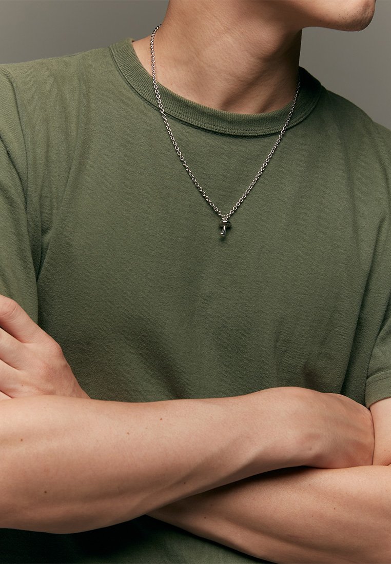 Vihreä oliivinvärinen pyöreä kaula-aukkoinen t-paita rentoa istuvuutta, yhdistettynä hopeaketjuun, jossa on pieni riipus. Kädet ristissä edessä.