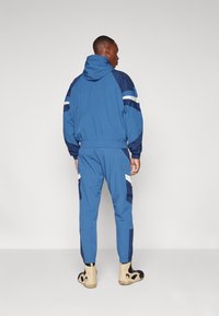 Blauwe trainingspak met een capuchon, voorzien van marineblauwe accenten en contrasterende witte strepen. Gemaakt van gladde stof, inclusief elastische manchetten en tailleband.
