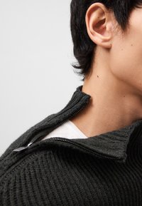 Maglione lavorato a maglia verde scuro con colletto alto, dotato di chiusura mezzi zip, texture a costine e indossato sopra una camicia bianca sottostante.