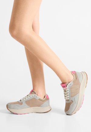 Nohy v béžových a sivých teniskách s ružovými akcentmi, jedna noha položená na zemi, druhá zdvihnutá, na jednoduchom bielom pozadí.