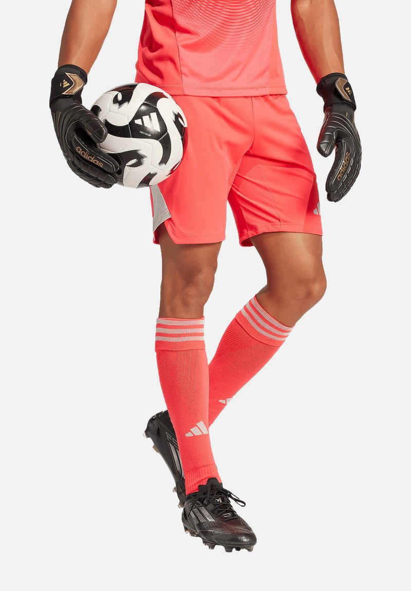 Rotes Sporttrikot mit horizontalen Streifen, passende Shorts, schwarze Handschuhe und schwarze Fußballschuhe. Hält einen schwarz-weißen Fußball.
