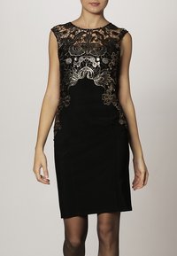 Svart klänning med figurnära silhuett, korta ärmar och intrikata guldspetsdetaljer runt halsringningen och bysten. Slät textur.