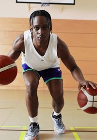 Jugador de baloncesto botando dos balones, vistiendo una camiseta sin mangas blanca, pantalones cortos morados con acentos verdes y zapatillas deportivas grises.