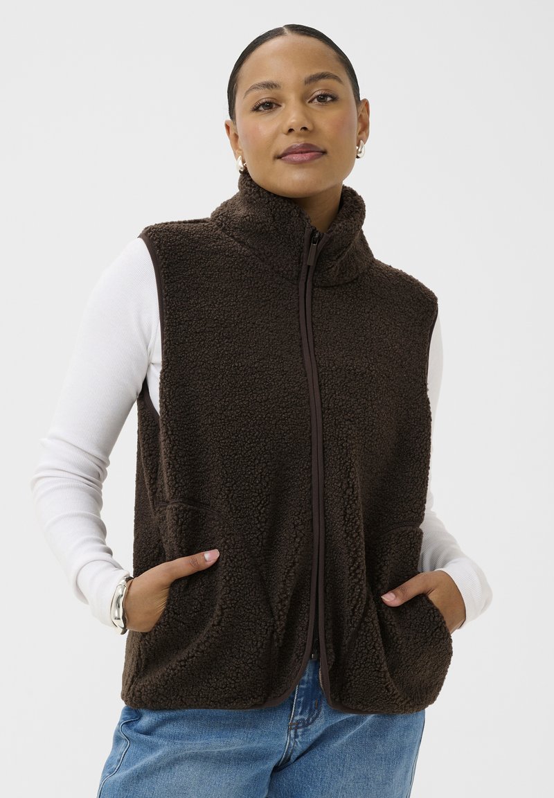 Mujer con chaleco de forro polar marrón oscuro con cremallera sobre camisa blanca de manga larga, con las manos en los bolsillos del chaleco, sobre un fondo liso.