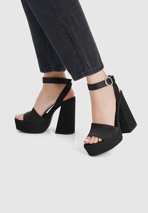 Person wearing black high-heeled platform sandals with ankle straps and black cropped pants against a plain background.