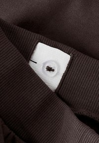 Brązowa dzianinowa tkanina swetra z prążkowanym kołnierzem. Biała metka z detalem guzika przy dekolcie. Miękka tekstura, casualowy design.