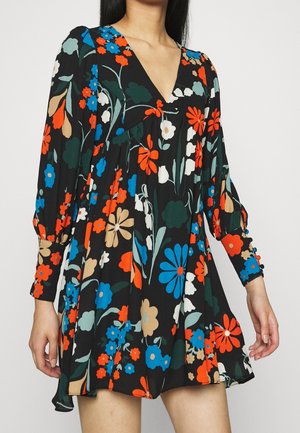 Vrouw die een zwarte bloemenjurk draagt met lange opgeblazen mouwen en levendige oranje, blauwe, beige, witte en groene bloemenpatronen.